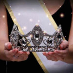 Sash-wearing person holding an ornate jeweled crown.