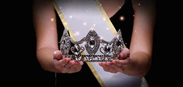Sash-wearing person holding an ornate jeweled crown.