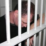 A man sitting in a jail cell with his head down, resting on the bars