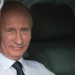 A smiling man in formal attire sitting inside a car with curtains
