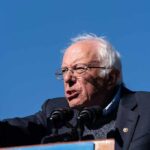 Bernie Sanders passionately speaking at a rally against a clear blue sky