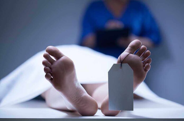 A deceased body on a table with a tag on the foot, and a medical professional in the background