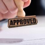 A hand holding a wooden stamp with the word 'APPROVED' above a document