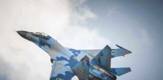 A blue camouflage fighter jet performing an aerial maneuver against a cloudy sky