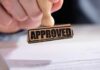 A hand holding a wooden stamp with the word 'APPROVED' above a document