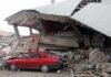 A red car parked next to a collapsed building with debris scattered around