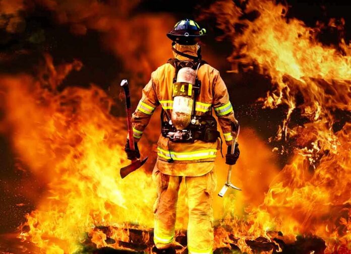 shutterstock_80081197 Firefighter standing in front of a large fire, equipped with tools