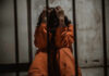 Person in orange jumpsuit sitting behind bars, handcuffed.