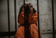 Person in orange jumpsuit sitting behind bars, handcuffed.
