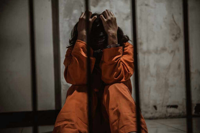 1484320151 Person in orange jumpsuit sitting behind bars, handcuffed.