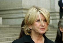 A woman with blonde hair in a black outfit, standing outdoors with a serious expression