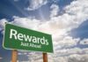 A green road sign indicating rewards just ahead against a cloudy sky