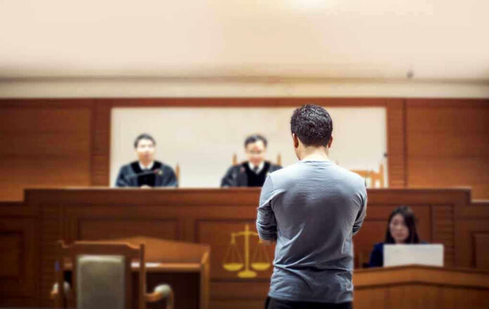 1049504825 Man stands in courtroom before judges and stenographer.
