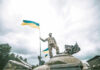 Russia Burns Through Men and Machines Soldier on tank holding Ukrainian flag.