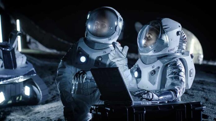 Two astronauts in spacesuits review data on a laptop on a moonlike surface at night
