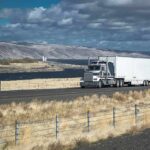 Semi-truck hauling a white trailer on a rural highway by a lake