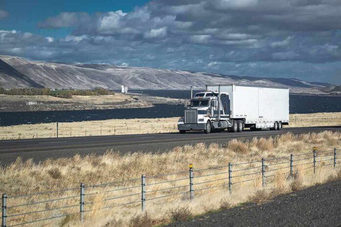 Semi-truck hauling a white trailer on a rural highway by a lake