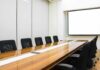 Conference room with long wooden table and empty chairs facing a blank projection screen