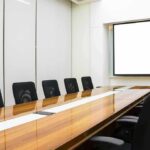 Conference room with long wooden table and empty chairs facing a blank projection screen