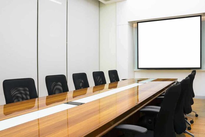 Conference room with long wooden table and empty chairs facing a blank projection screen