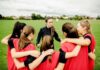 Backlash Explodes After Bold Locker-Room Stand Coach talks to youth team in a huddle on a field