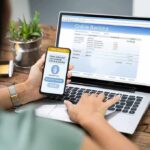 New Files Reveal FBI Surveillance Tactics Person using smartphone to approve bank transfer beside laptop with online banking