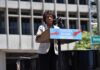 Maxine Waters speaks at an outdoor podium with a protest sign