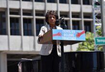 Maxine Waters speaks at an outdoor podium with a protest sign