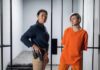 Police officer stands beside inmate in orange uniform inside a jail cell