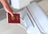 Hand holding a red passport next to a gray suitcase