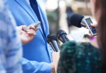 Reporters hold microphones toward a person in a blue suit