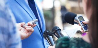 Reporters hold microphones toward a person in a blue suit